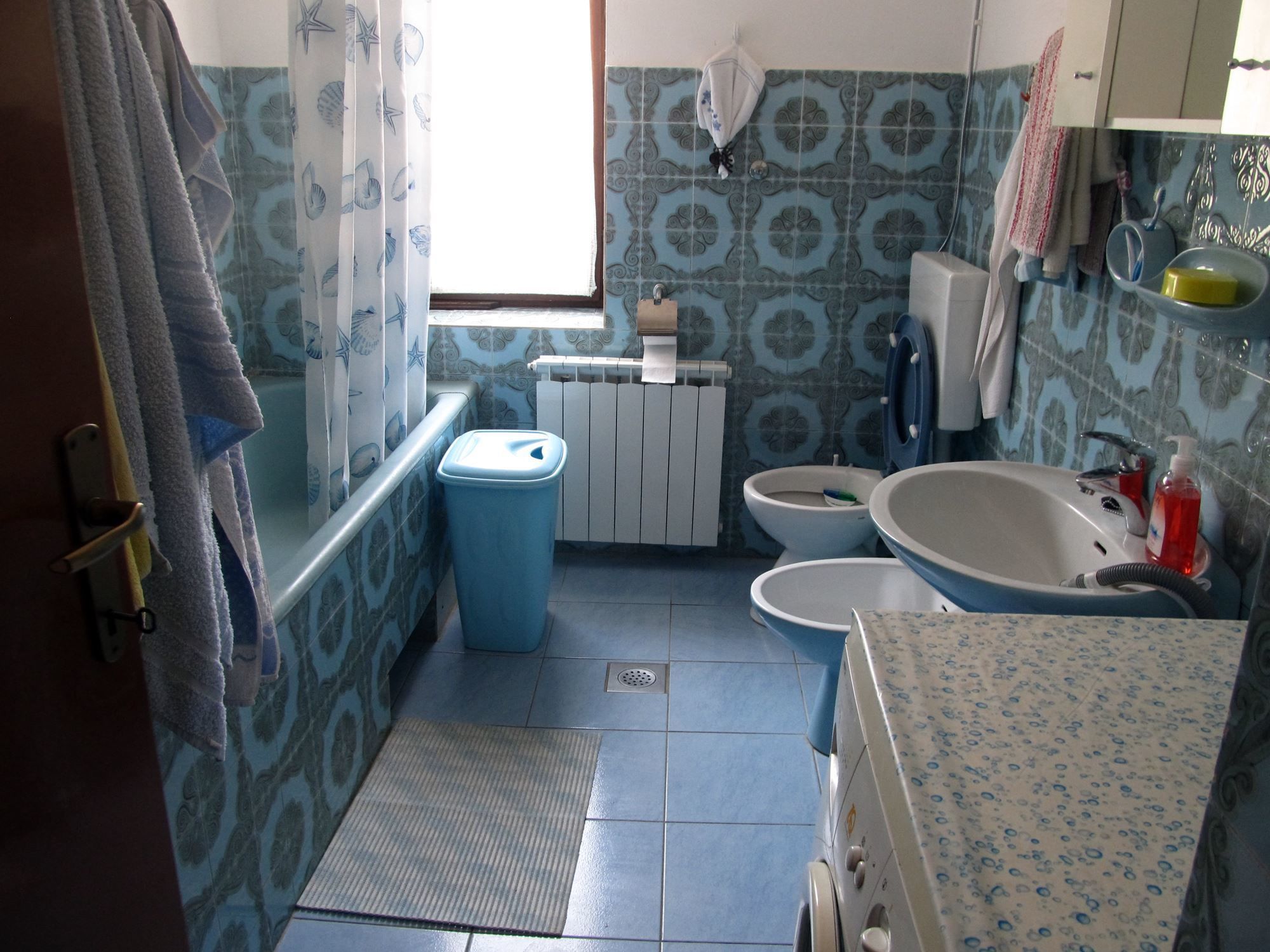 Ein Badezimmer mit Toilette, Bidet und Badewanne. Ein Fenster mit Vorhängen, ein Handtuchhalter und ein Mülleimer sind vorhanden.