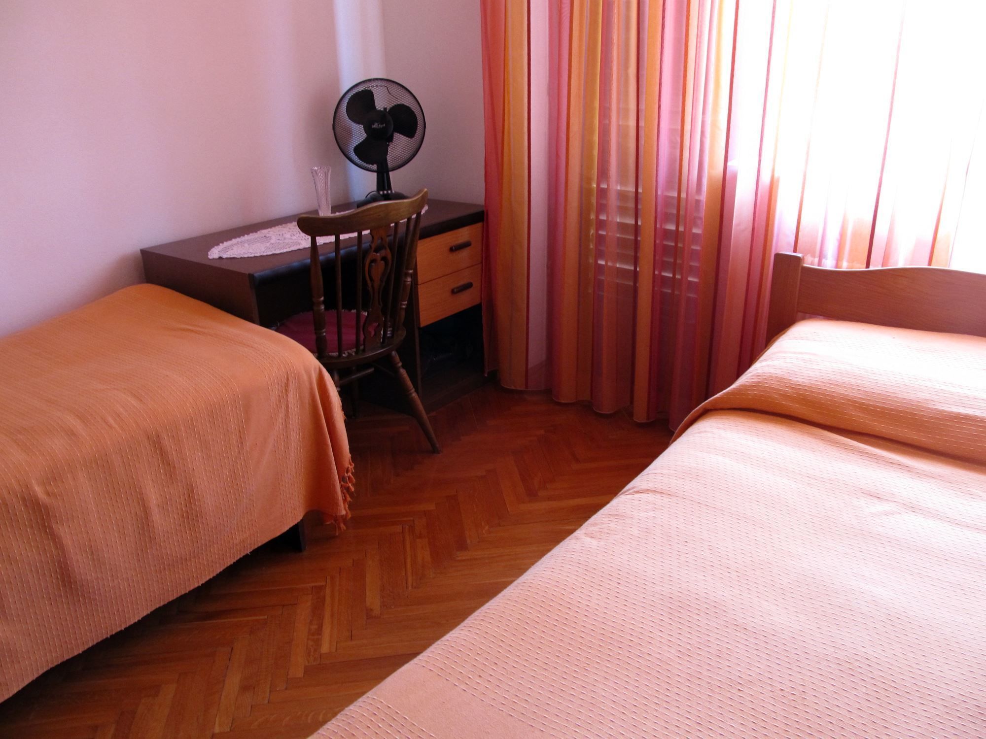 Ein Zimmer mit zwei Betten, einem Schreibtisch mit Stuhl, einem Ventilator und einem Fenster mit Vorhängen.