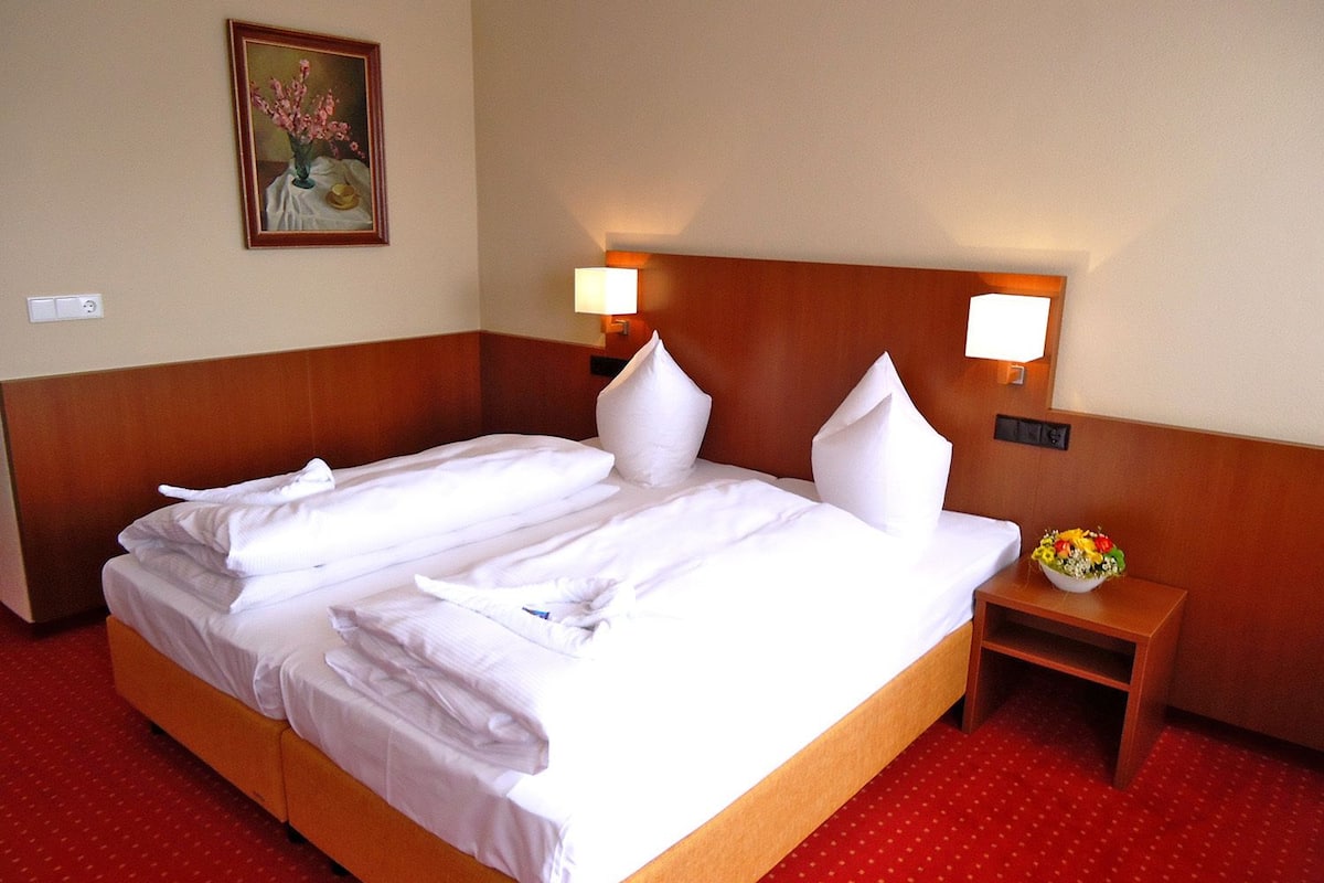 A hotel room with a large bed, two bedside lamps, a small table with a flower arrangement, and a painting on the wall.