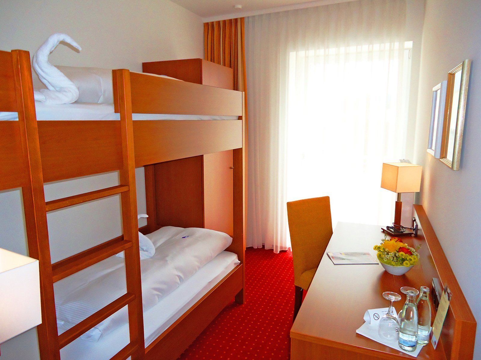 Ein Hotelzimmer mit Etagenbett, Schreibtisch mit Lampe, Stuhl, kleinem Tisch mit Blumenvase und einem Fenster mit Vorhängen.