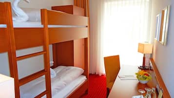 Ein Hotelzimmer mit Etagenbett, Schreibtisch mit Lampe, Stuhl, kleinem Tisch mit Blumenvase und einem Fenster mit Vorhängen.