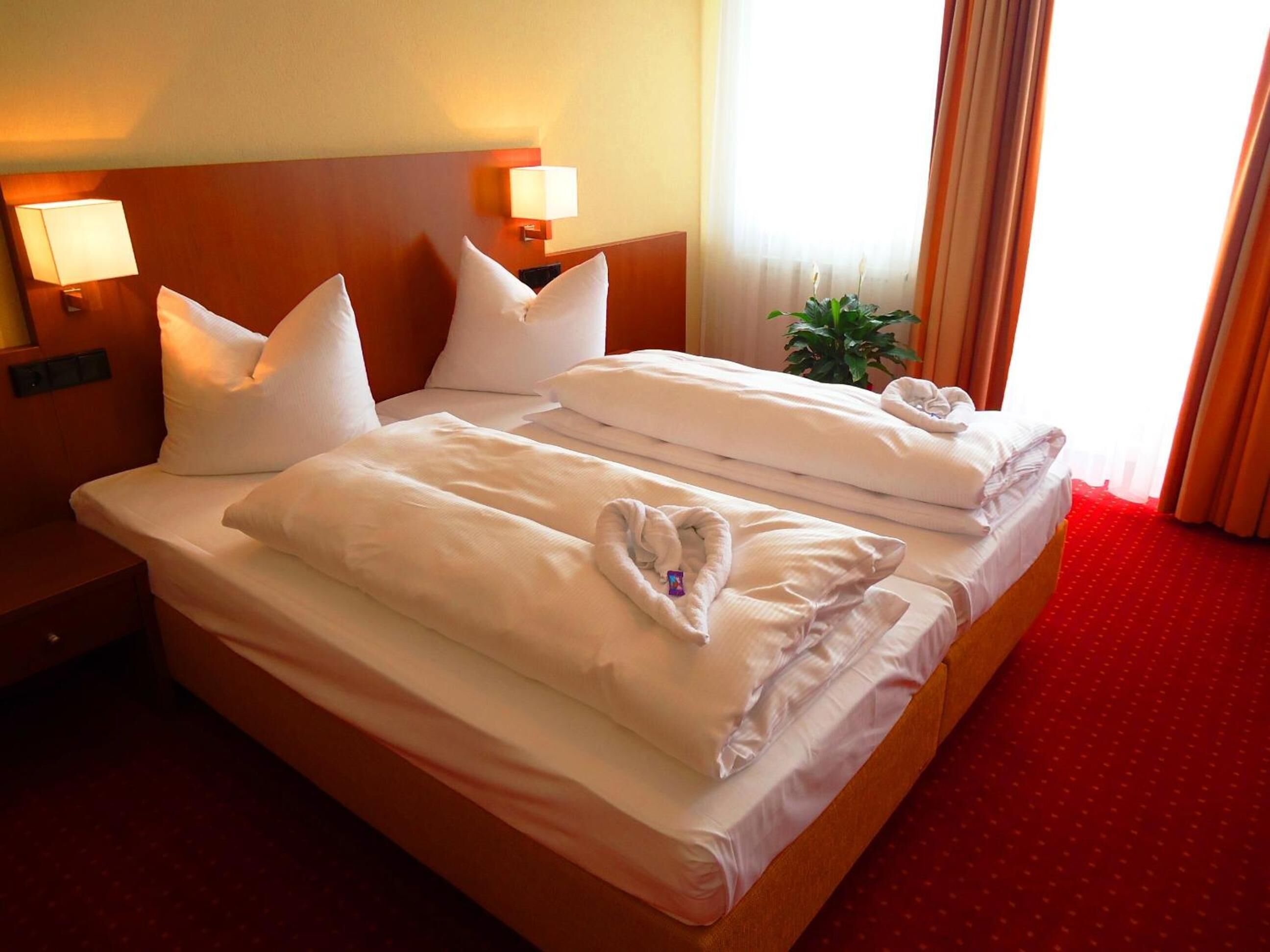 A double bed with white linens and pillows, two bedside lamps, a red carpeted floor, and a window with curtains.
