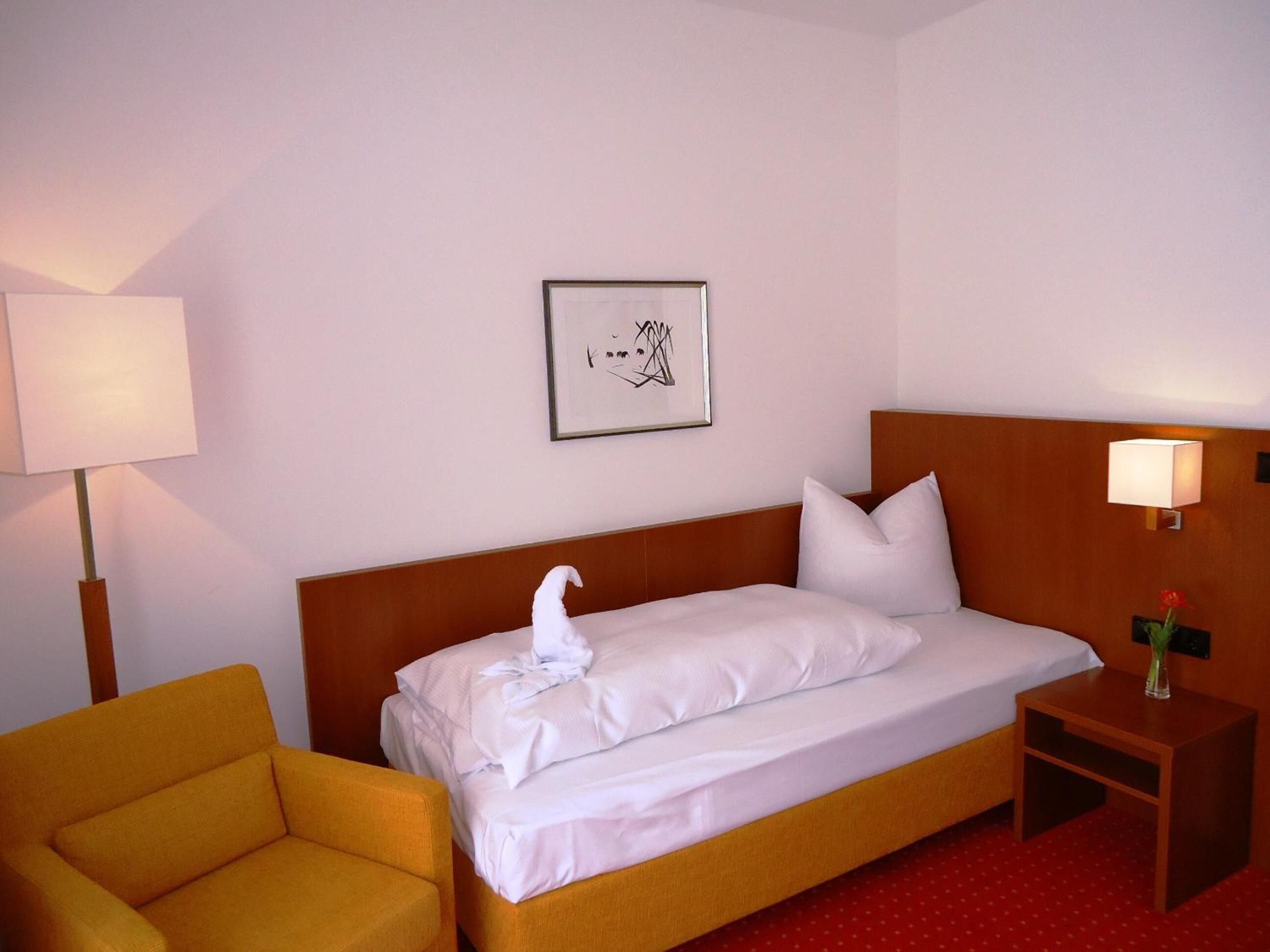 Una habitación de hotel con una cama, una silla, una mesita de noche con lámpara y un cuadro en la pared.