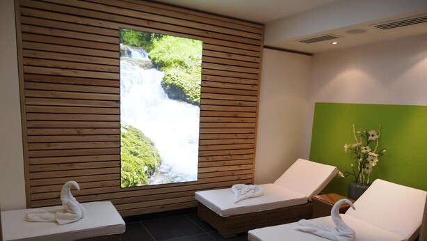A spa room with three lounge chairs, each with a towel and a swan-shaped towel on the headboard. A large window displays a waterfall view. - Hotel Gasthof Posthalter (Zwiesel)
