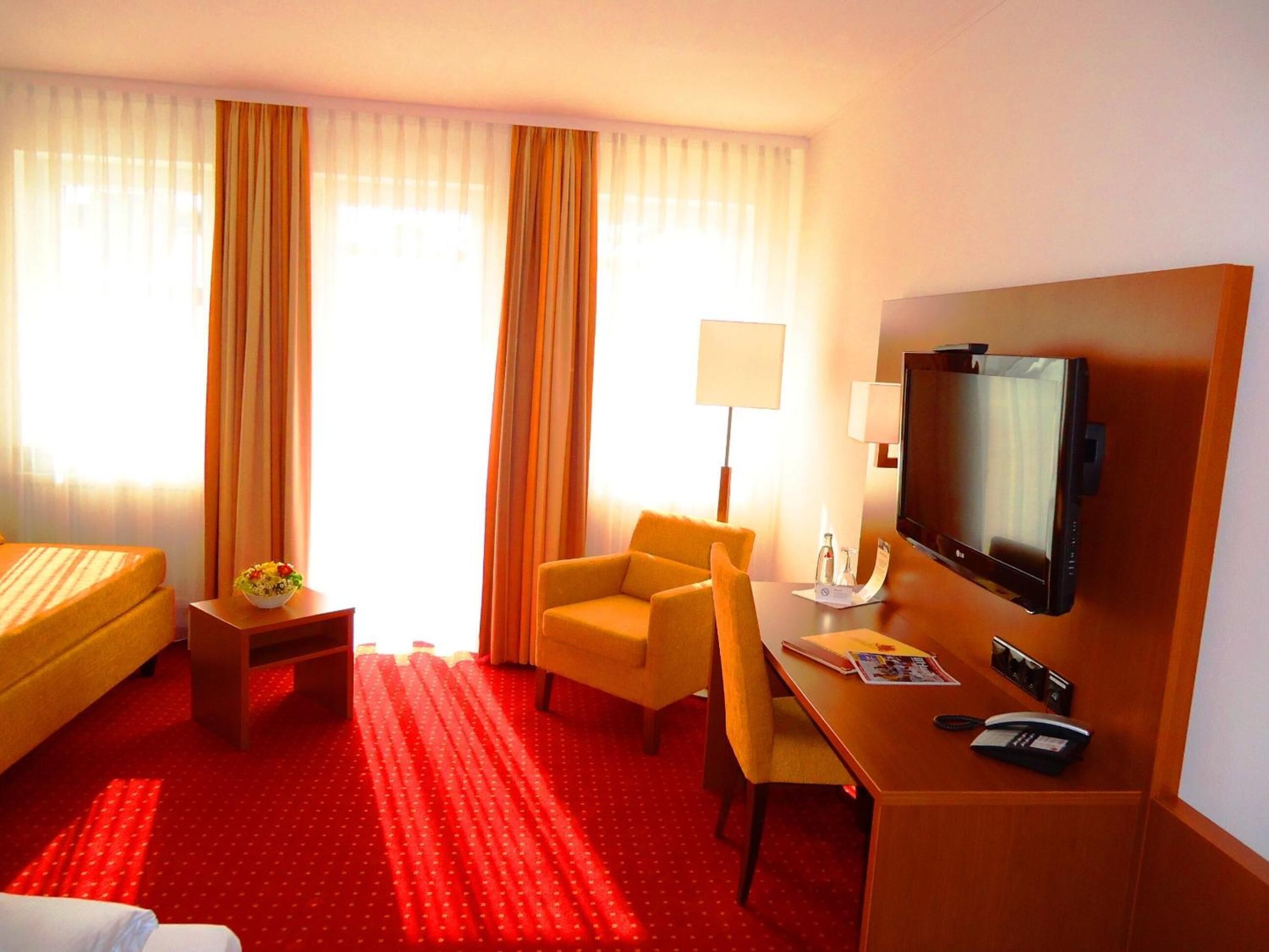 Quarto de hotel com cama, escrivaninha com cadeira, televisão e luminária.