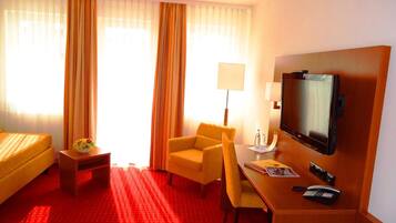 Quarto de hotel com cama, escrivaninha com cadeira, televisão e luminária.