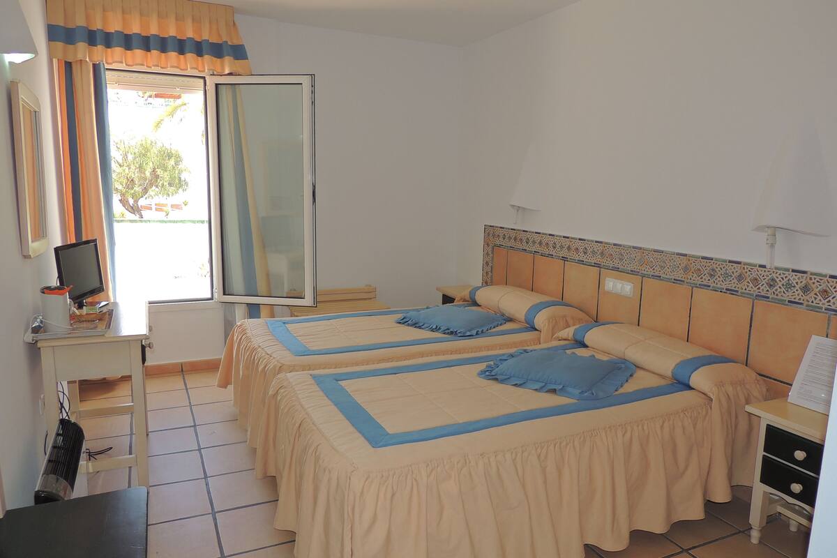 Ein Hotelzimmer mit zwei Betten, einem Schreibtisch mit Computer und einem Fenster mit Vorhängen.