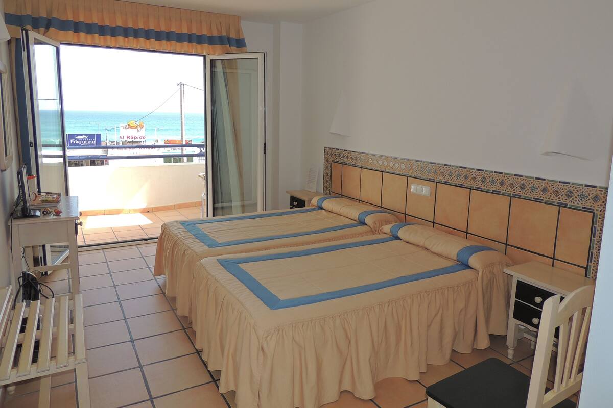 Ein Hotelzimmer mit zwei Betten, einem Schreibtisch und einem Stuhl. Es gibt einen Balkon mit Blick auf das Meer und ein Gebäude mit einem Schild.
