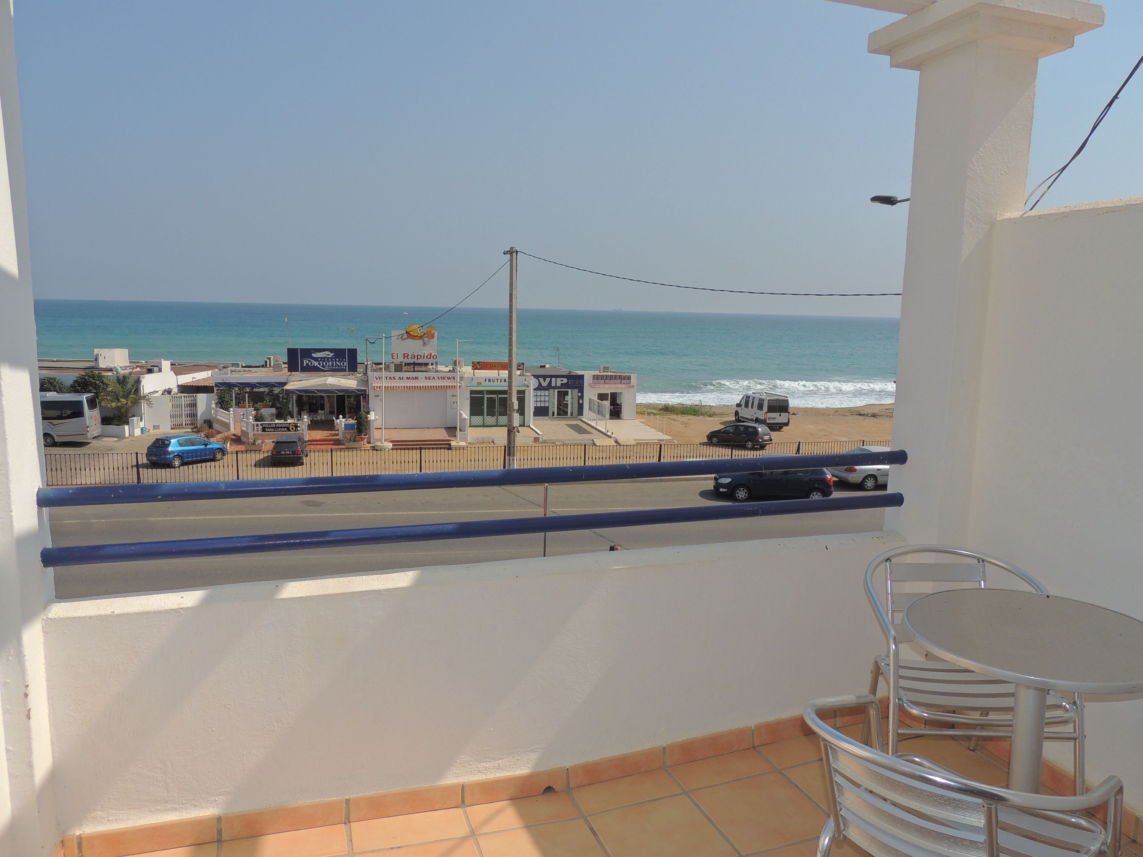A balcony with a view of the beach, a road with parked cars, and a few buildings including a restaurant named "El Rapido".