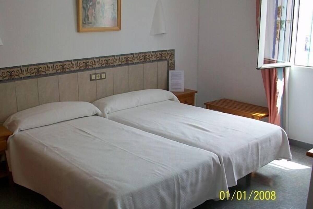 Ein Doppelbett mit weißen Bettwäsche in einem Hotelzimmer. Es gibt zwei Nachttische, jeweils einer an den Seiten des Bettes. Über dem Bett hängt ein Gemälde, und zur Rechten ist ein Fenster mit Vorhängen zu sehen.