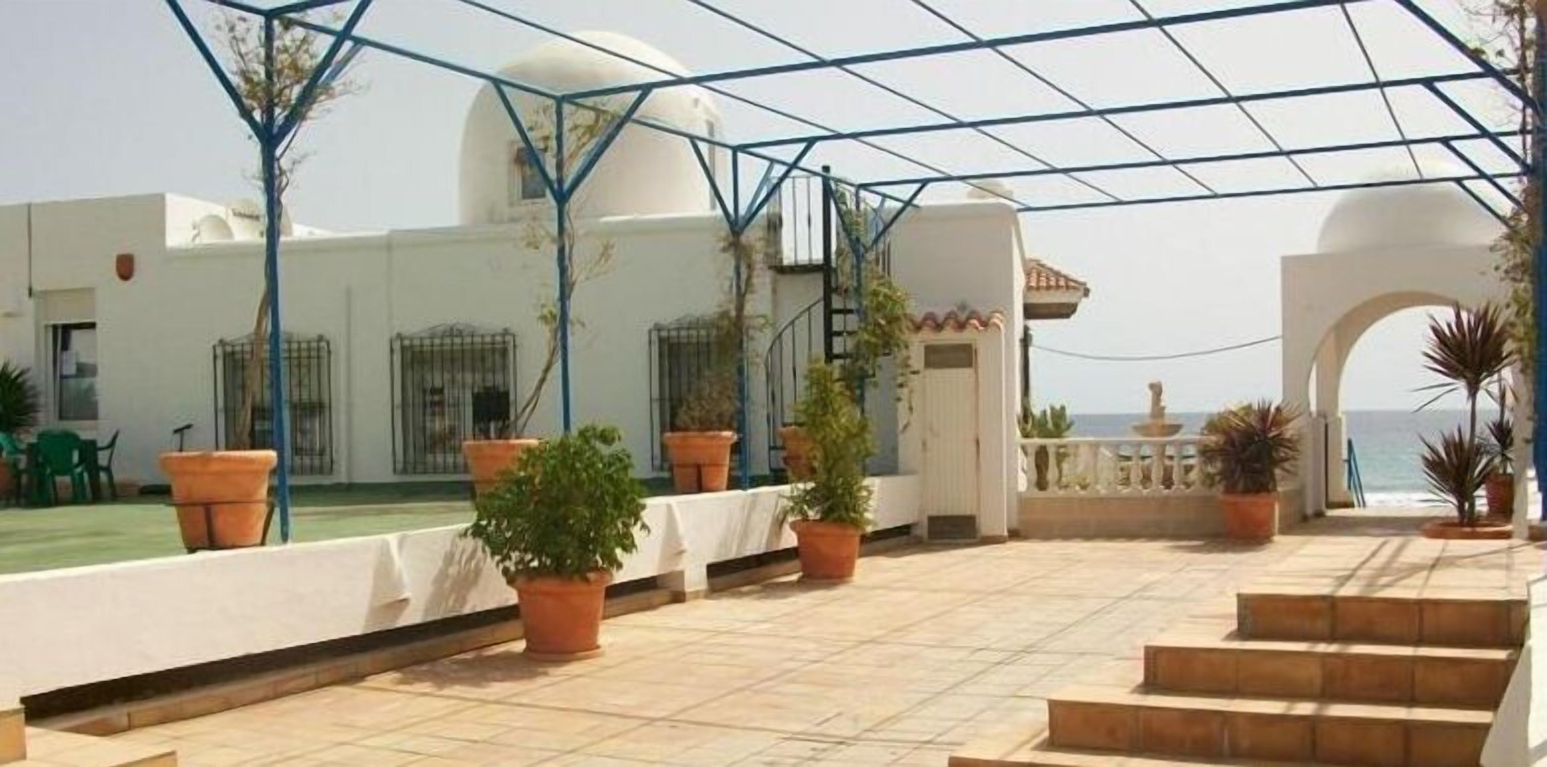 Un espace extérieur abrité, agrémenté de plantes en pot, un bâtiment blanc surmonté d’un dôme et une vue sur la mer.
