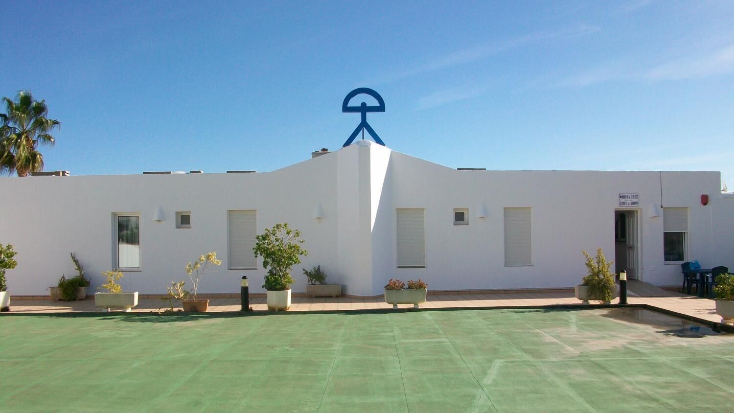 Un edificio blanco con un símbolo azul en el techo, plantas en macetas y un área verde al aire libre.