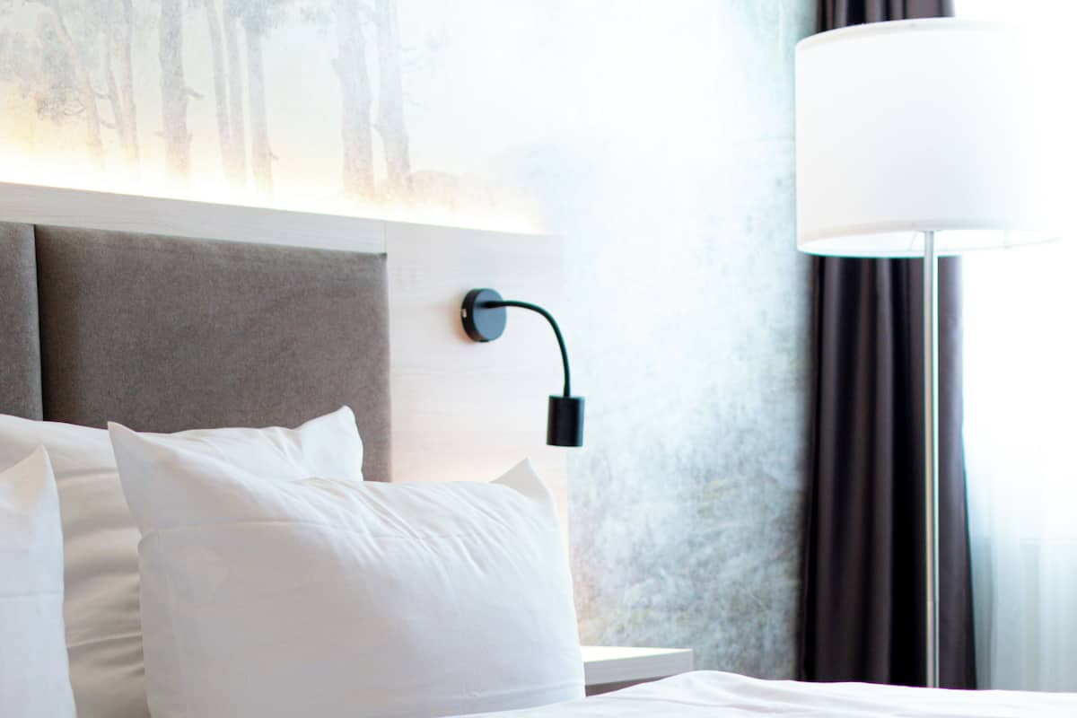Un lit bien fait, avec du linge de lit blanc, une tête de lit et une lampe fixée au mur.