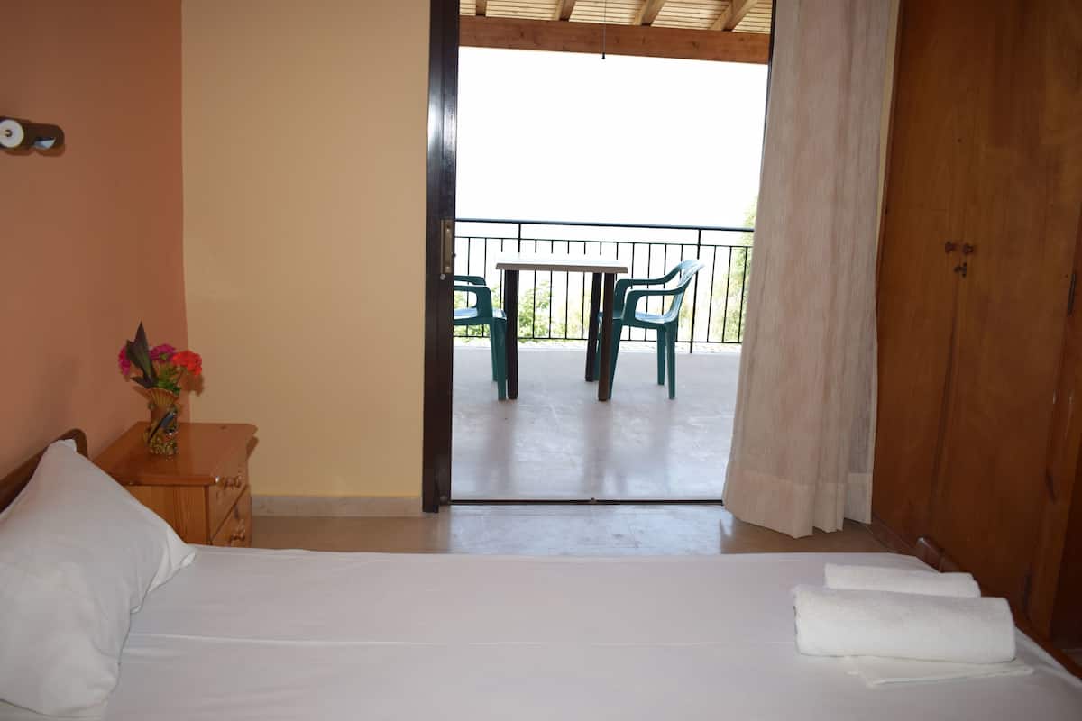 A hotel room with a bed, a nightstand with a vase of flowers, a balcony with a table and chairs, and a wooden door.