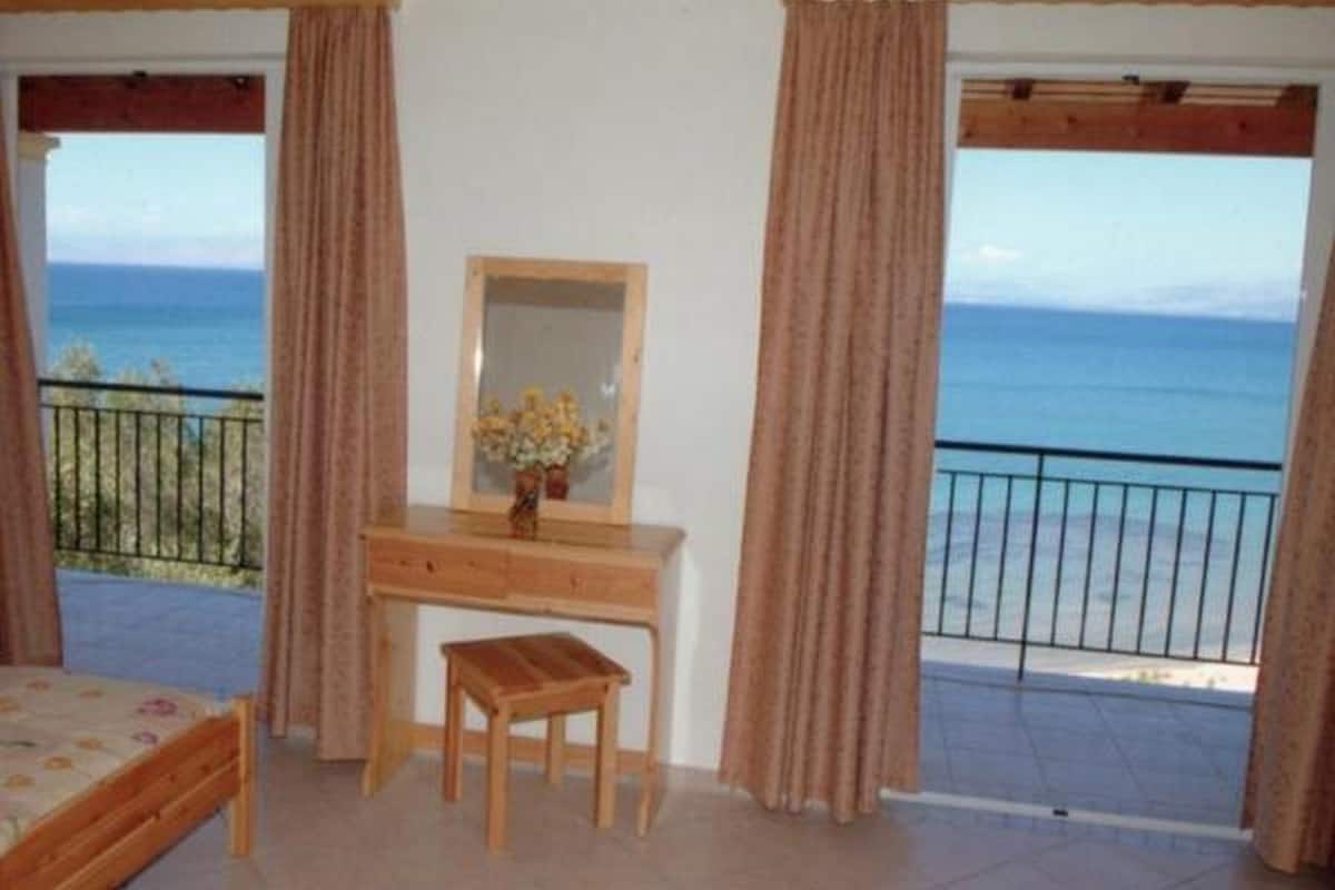 A bedroom with a wooden desk, a small stool, a mirror, and a view of the sea.