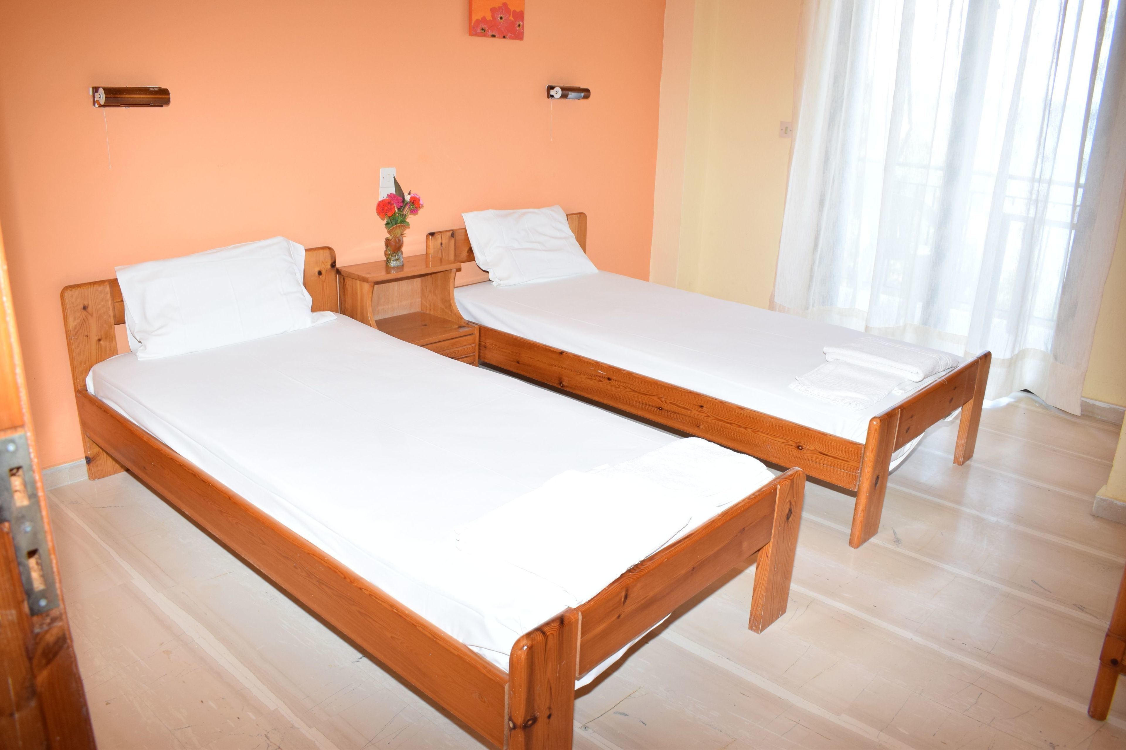 A room with two wooden beds, a nightstand with a vase of flowers, and a window with sheer curtains.