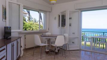 Una stanza con un tavolo da pranzo, sedie, una televisione su un mobile, un termosifone e vista sul mare attraverso porte aperte.