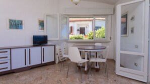 Una stanza con un tavolo da pranzo, sedie, una televisione su un mobile e una finestra vista su un edificio e vegetazione.