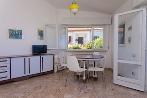 A room with a dining table, chairs, a television on a cabinet, and a window with a view of a building and greenery.