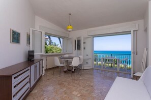 A room with a dining area, a TV on a cabinet, a balcony with a view of the sea, and a white sofa.