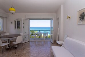 A room with a dining table, chairs, a white sofa, a yellow pendant light, and a view of the sea through an open door.