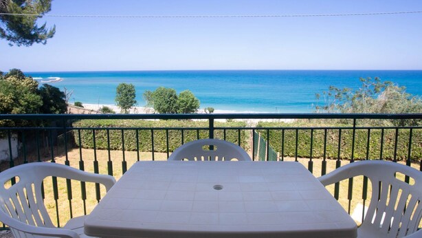 Un balcone con tavolo e sedie, vista su un mare cristallino e una spiaggia.