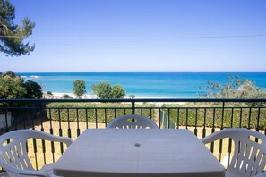 A balcony with a table and chairs, overlooking a clear blue sea and a beach.