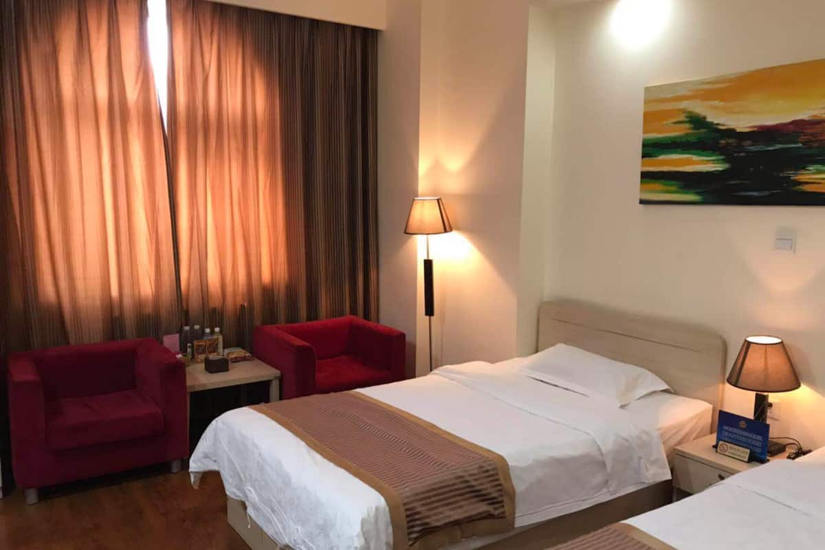 A hotel room with two beds, a red chair, a nightstand with a lamp, and a painting on the wall.