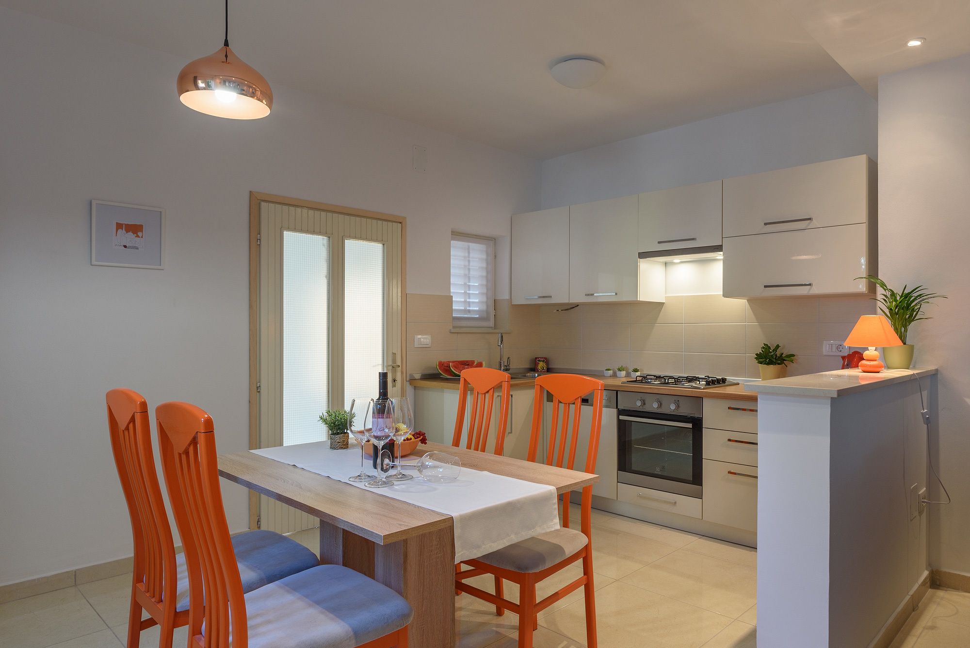 A compact kitchen with a dining area, featuring a wooden table, orange chairs, and a small island with a stove.