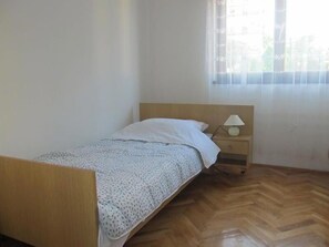 Un petit lit bien fait, avec une tête de lit en bois, une table de chevet avec une lampe et une fenêtre ornée de rideaux légers.