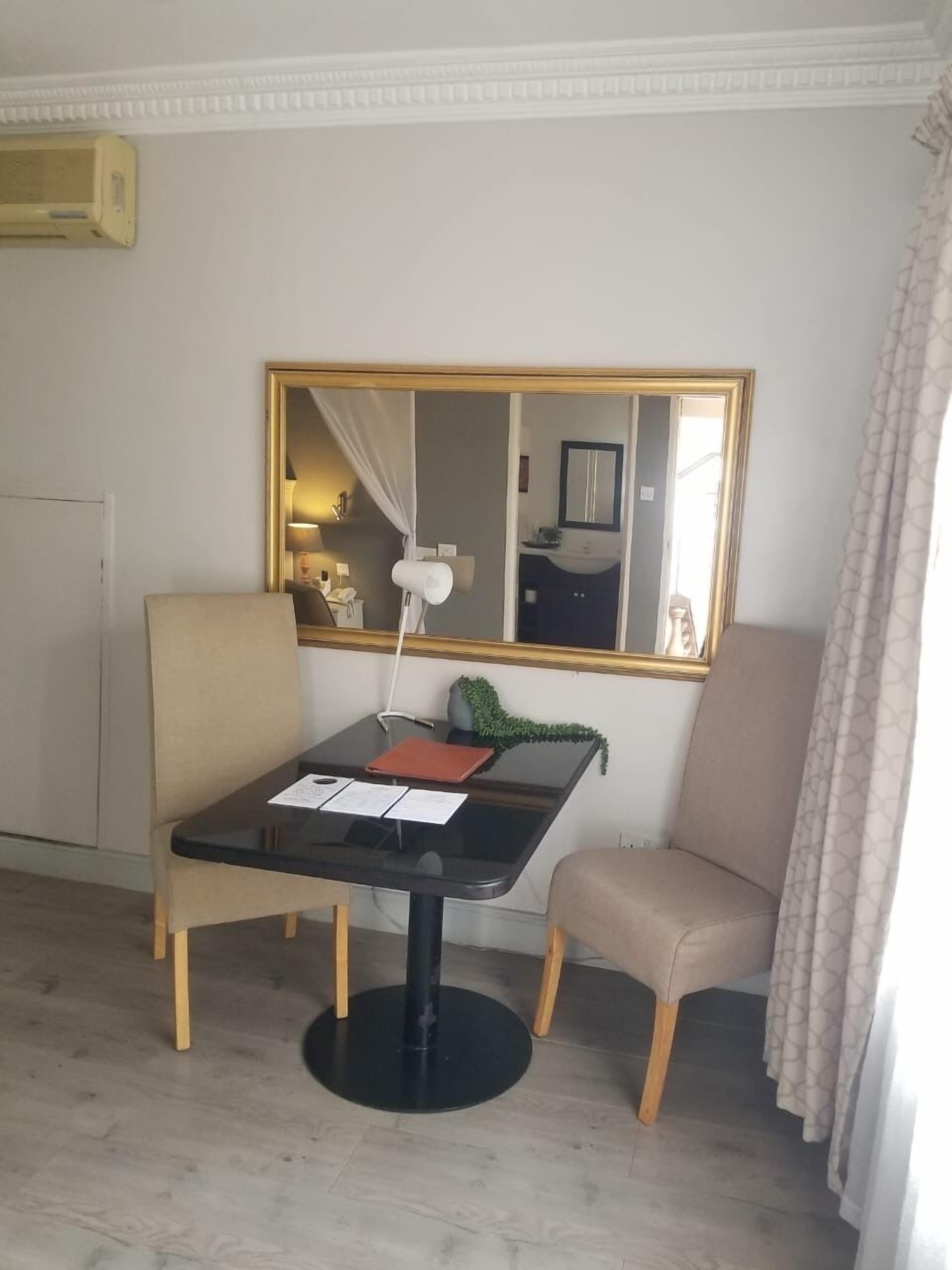 Ein kleiner, gut beleuchteter Raum mit einem runden Tisch, zwei Stühlen, einem Spiegel und einem Fenster mit Vorhängen.