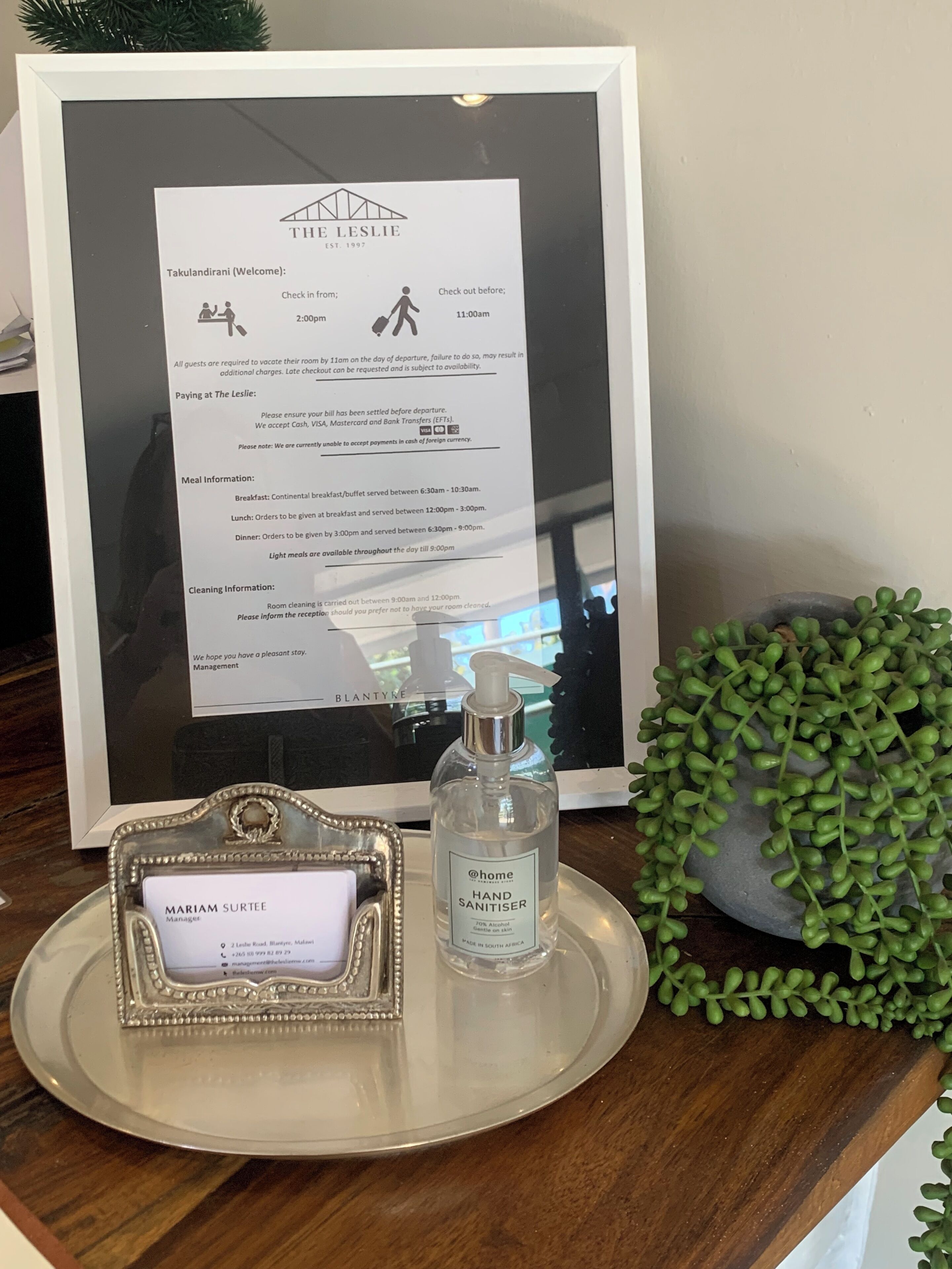 A framed welcome sign with check-in and check-out times, a hand sanitizer bottle, and a nameplate for the manager.