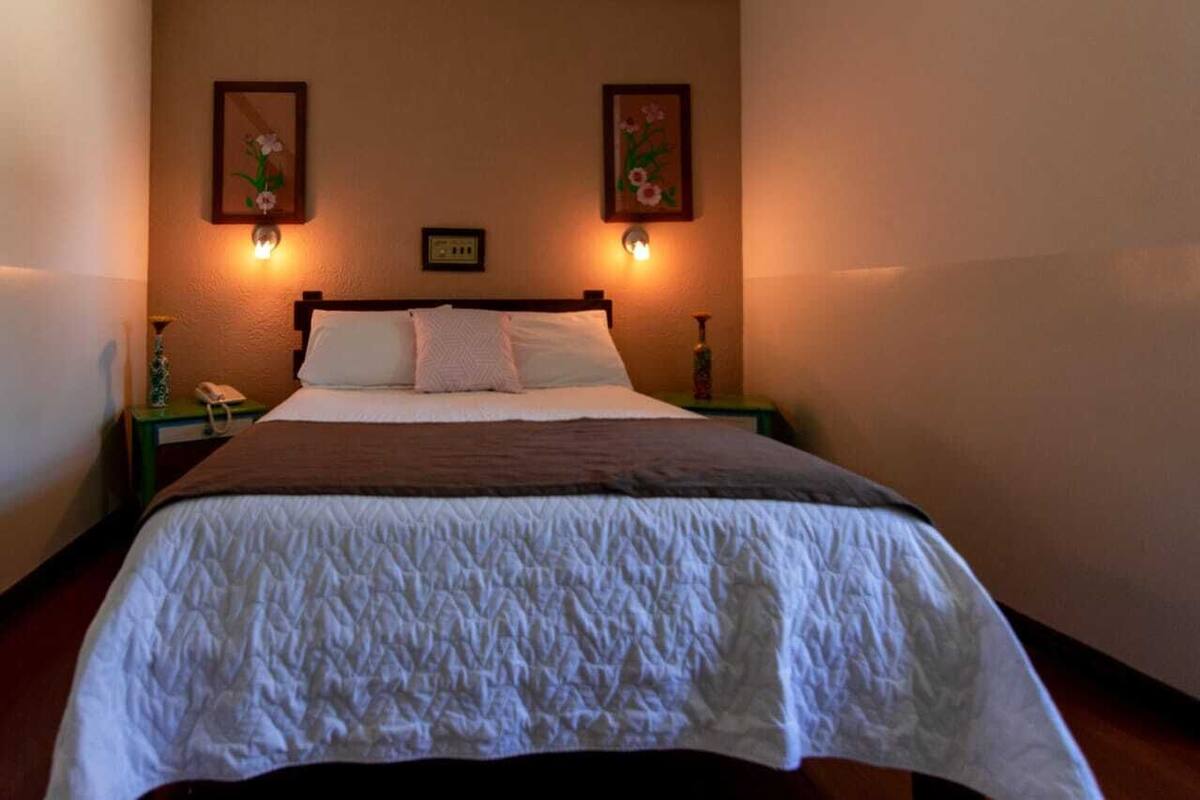 Una cama bien hecha con colcha blanca, dos lámparas de pared y dos cuadros enmarcados con motivos florales encima.