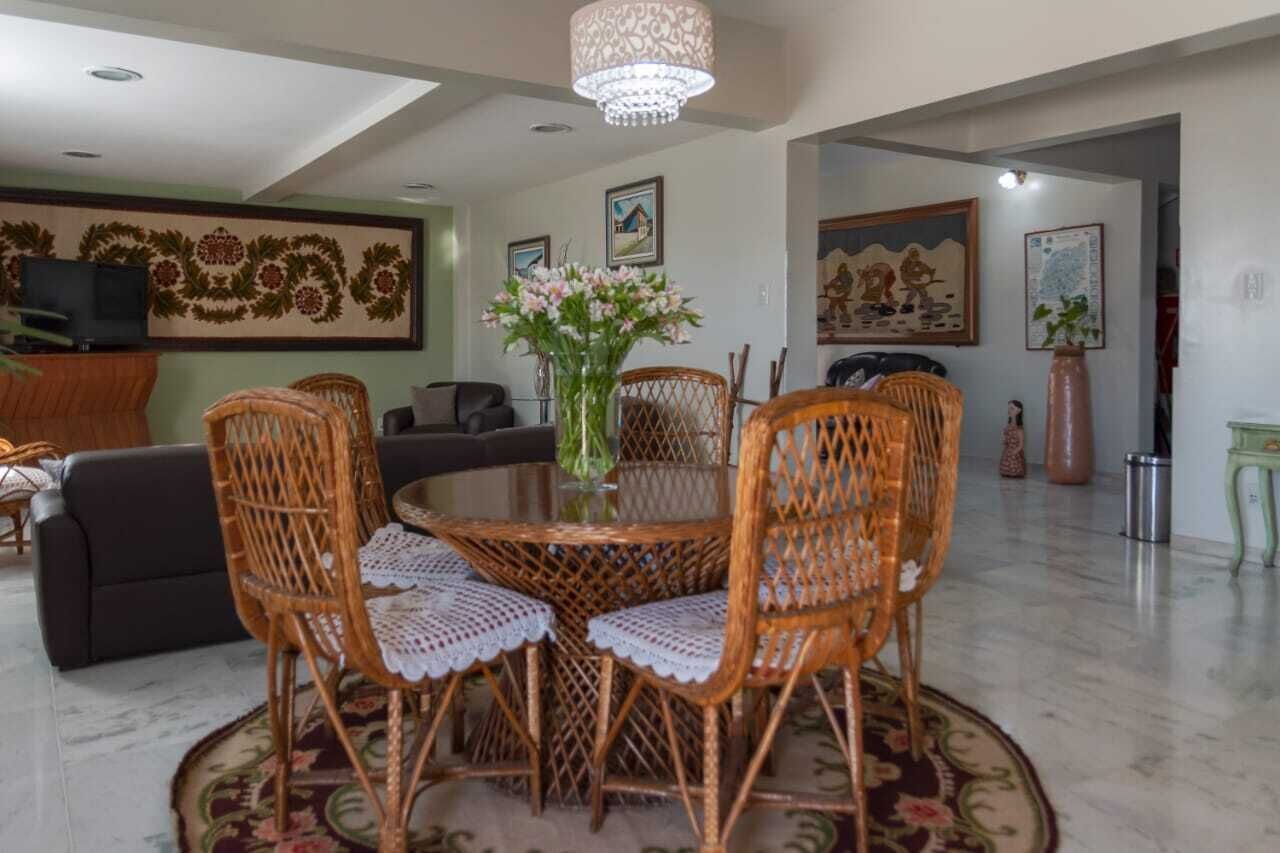 A dining area with a round table surrounded by wicker chairs, a chandelier, and a vase with flowers.