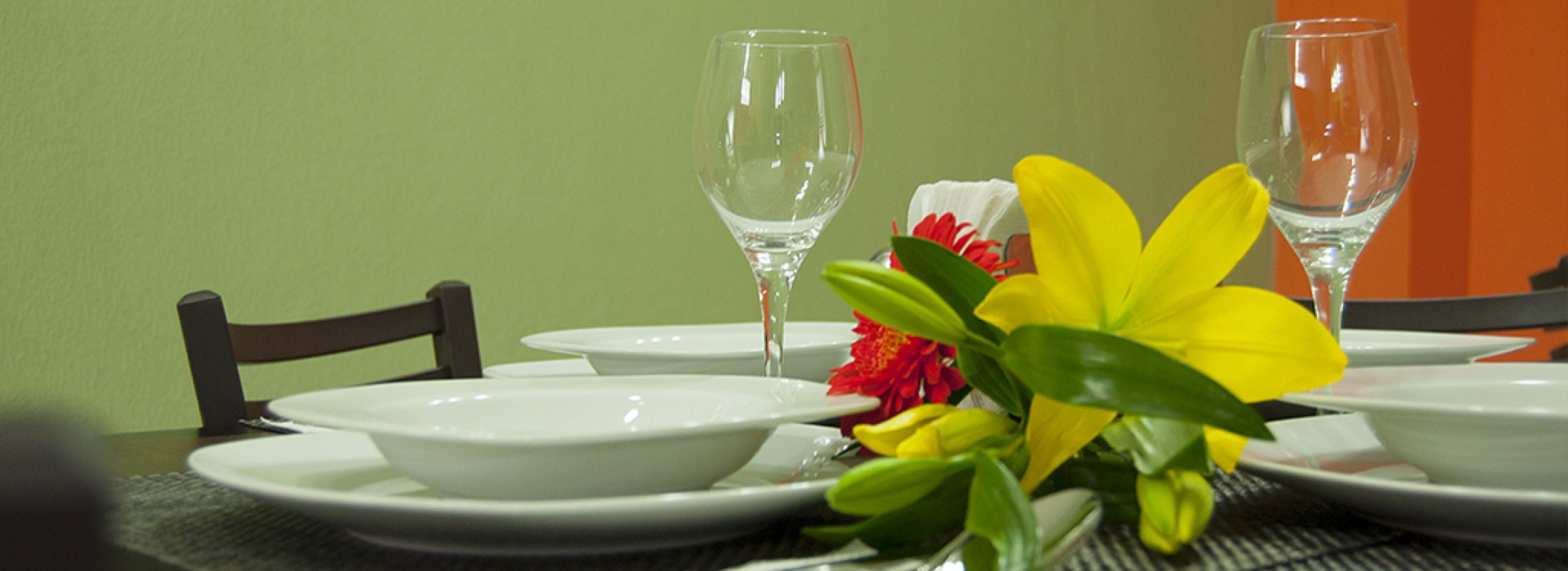 Una mesa dispuesta con platos, tazones y vasos blancos, junto a un arreglo floral amarillo.