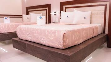 Habitación de hotel con dos camas, cada una con cabecero de madera y colcha estampada. Hay dos mesitas de noche con lámparas y una luz en la pared.