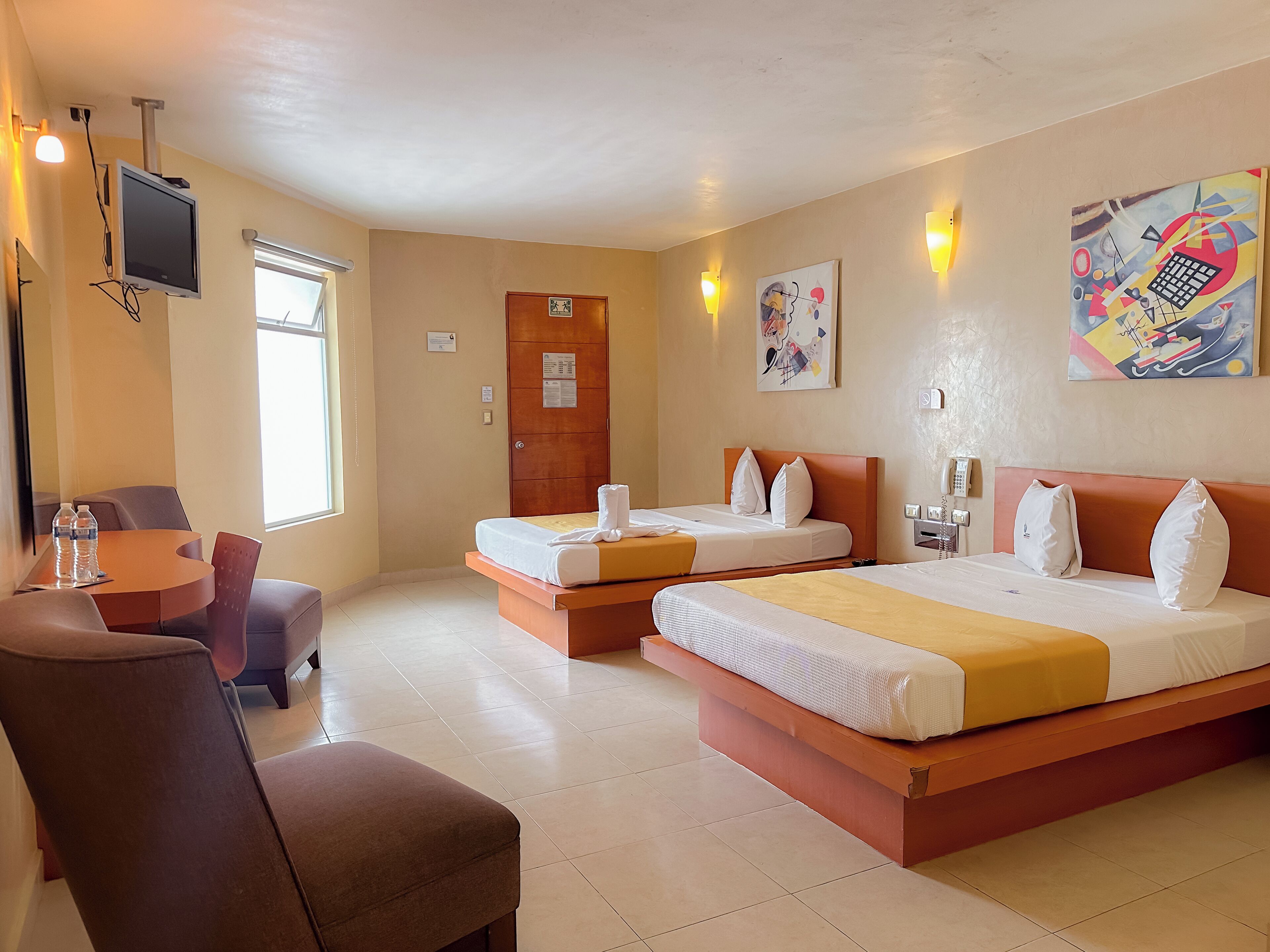 Habitación de hotel con dos camas, un escritorio y sillas, televisor y obras de arte en las paredes.
