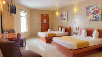 Quarto de hotel com duas camas, uma escrivaninha com cadeiras, televisão e obras de arte nas paredes.