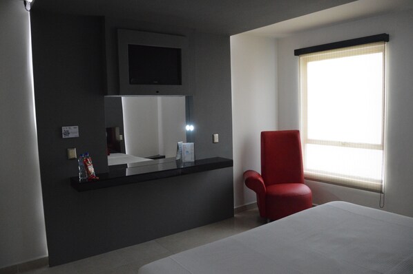 Habitación de hotel con cama, una silla roja, mostrador de recepción, televisor y ventana con persianas.