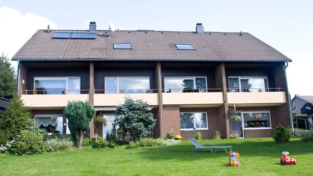 A two-story residential building with a flat roof, multiple windows, and a small garden area with a lawn, a tree, and outdoor furniture. - FeWo Walter - House 2 St.- Andreasberg, south-facing slope, quiet, family-friendly, WLAN (St. Andreasberg)