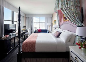 A hotel room with a large bed, a desk with a TV, a chair, and a view of the city.