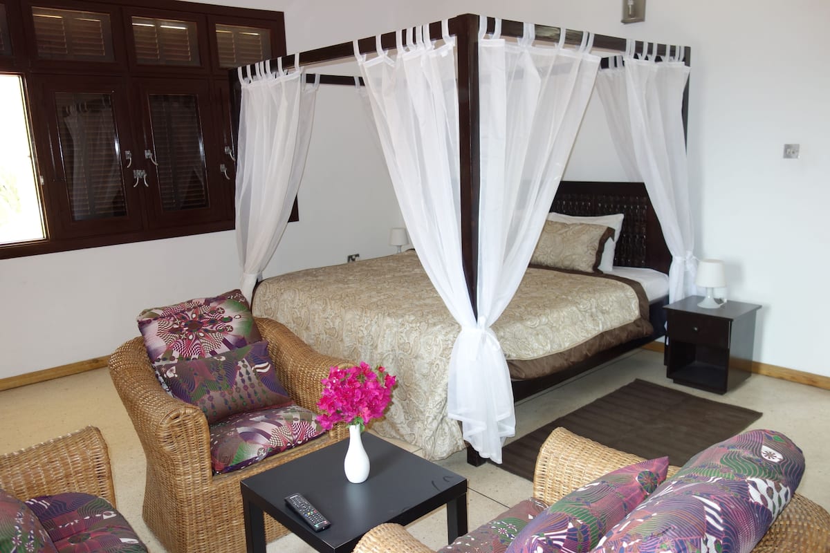 A bedroom with a four-poster bed, a sitting area with a sofa and armchair, a small table with a vase of flowers, and a nightstand with a lamp.