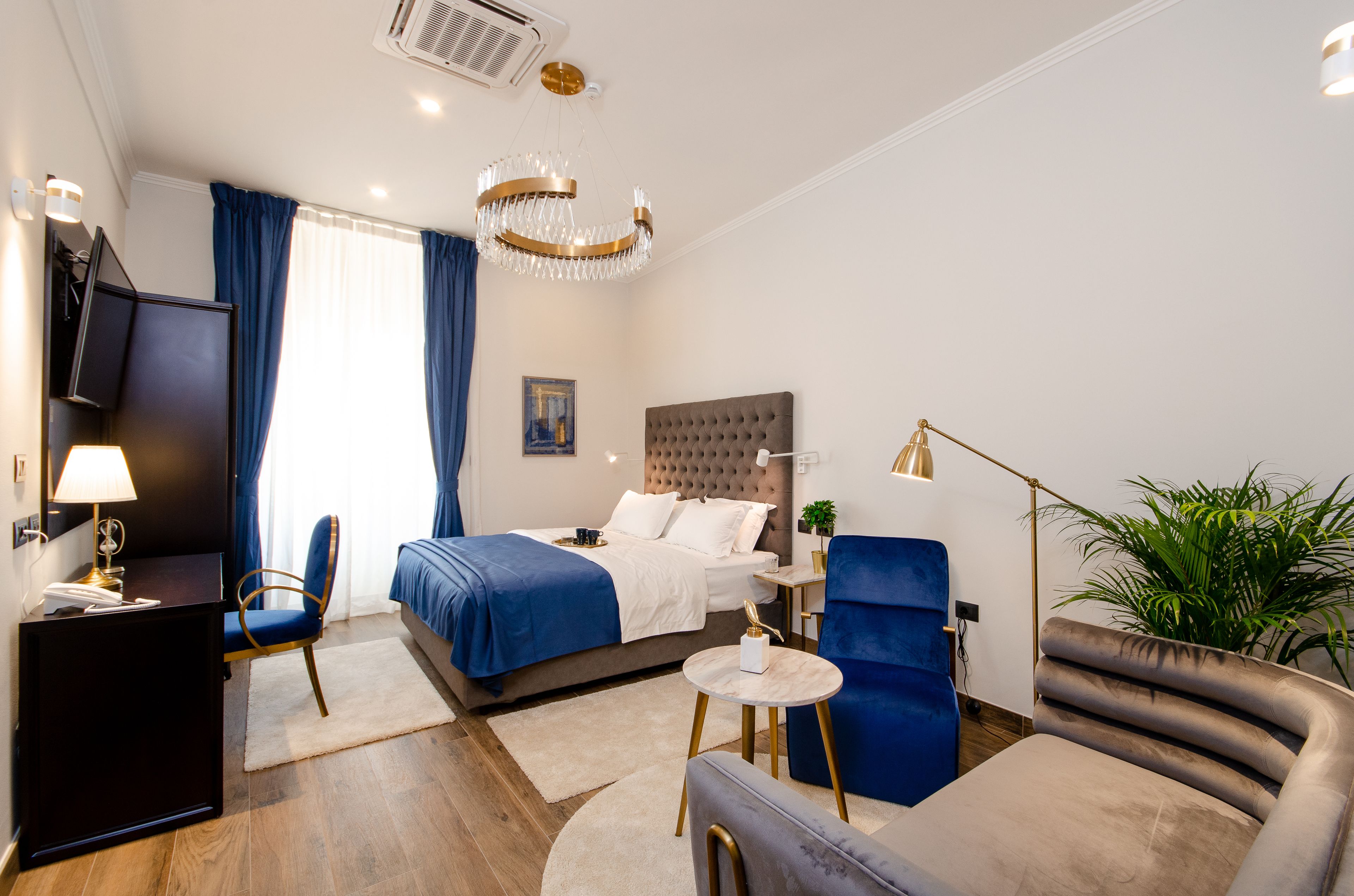 A modern hotel room with a bed, a desk, a chair, a sofa, and a potted plant.