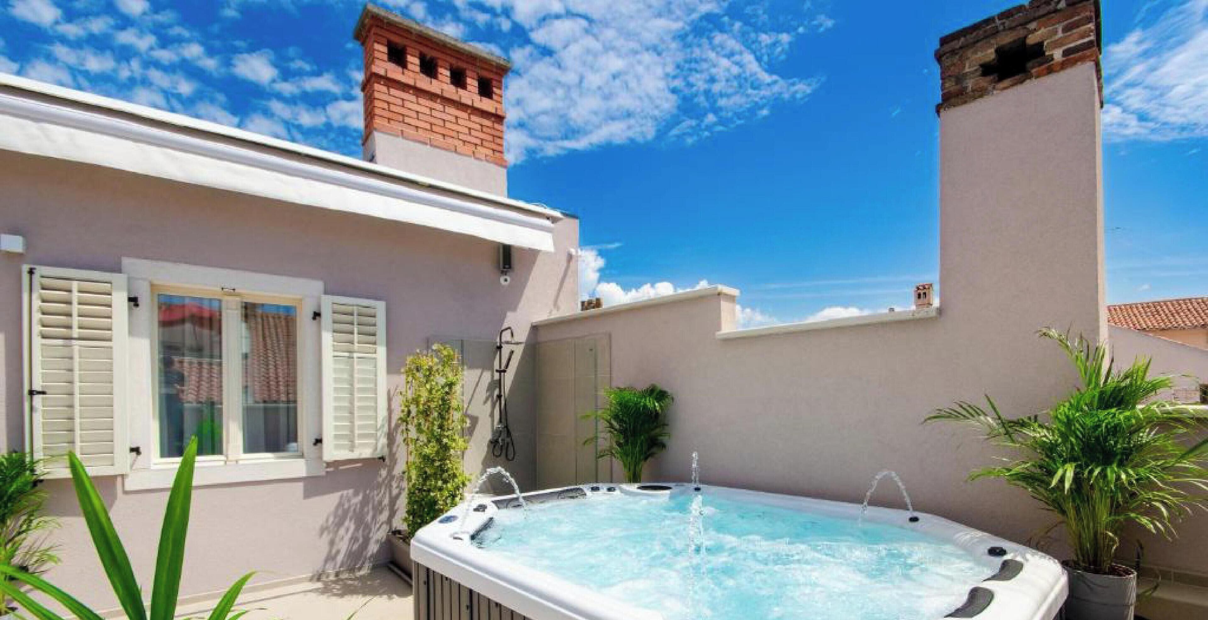 Un bain à remous dans un jardin, avec une maison, une cheminée en briques et des plantes en pot.