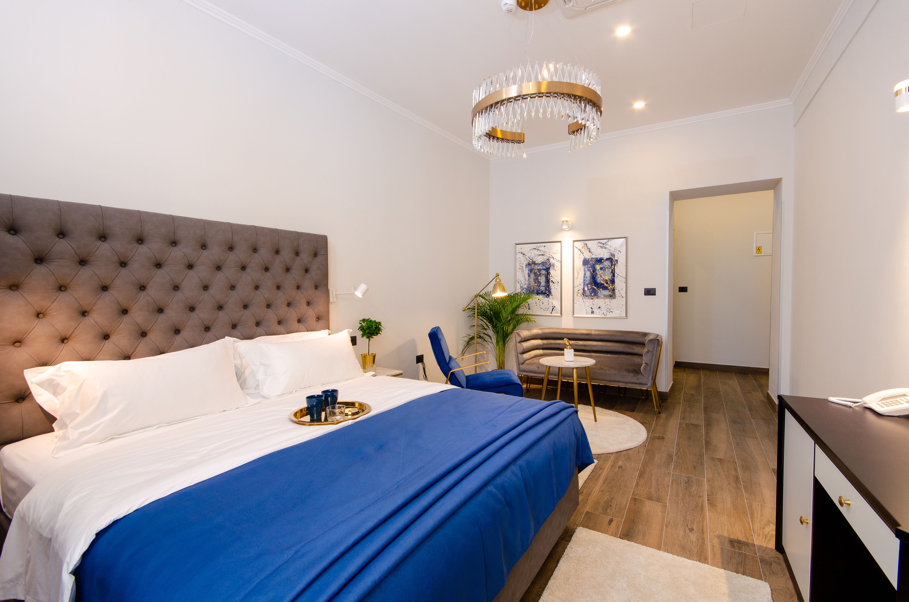 A hotel room with a tufted headboard bed, a blue bedspread, a chandelier, a desk with a telephone, and a small round table with a plant.