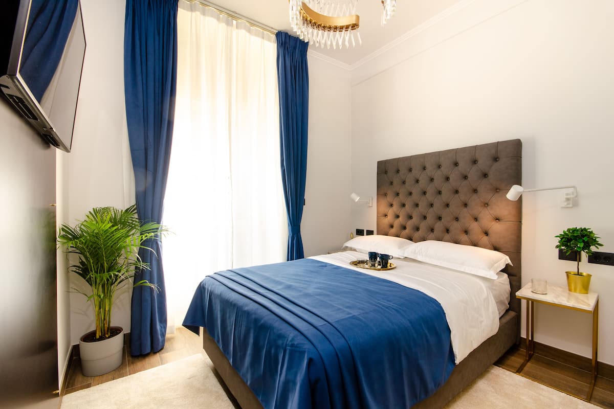 Una cama bien hecha con un edredón azul, una planta en maceta y una mesita de noche con una lámpara y una pequeña planta.