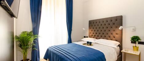 A neatly made bed with a blue bedspread, a potted plant, and a bedside table with a lamp and a small plant.