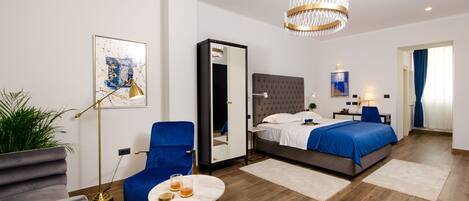 A modern hotel room with a large bed, a blue armchair, a round table with a drink, a mirror, and a chandelier.