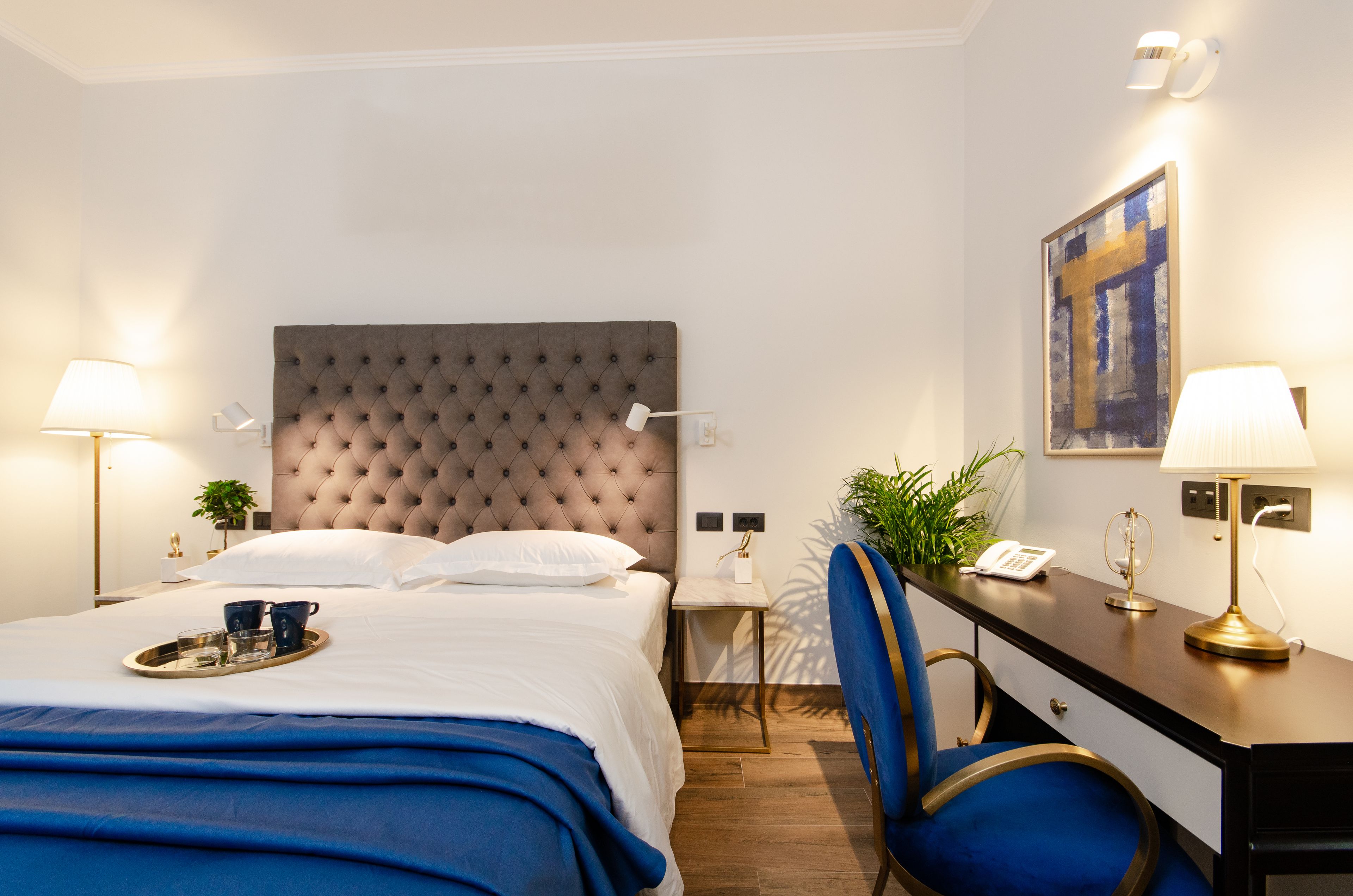 Ein Hotelzimmer mit einer gebl ümten Kopfteil, einem Schreibtisch mit Telefon, einem Sessel und einem Nachttisch mit Lampe sowie einer Ablage mit Tassen und Gläsern.