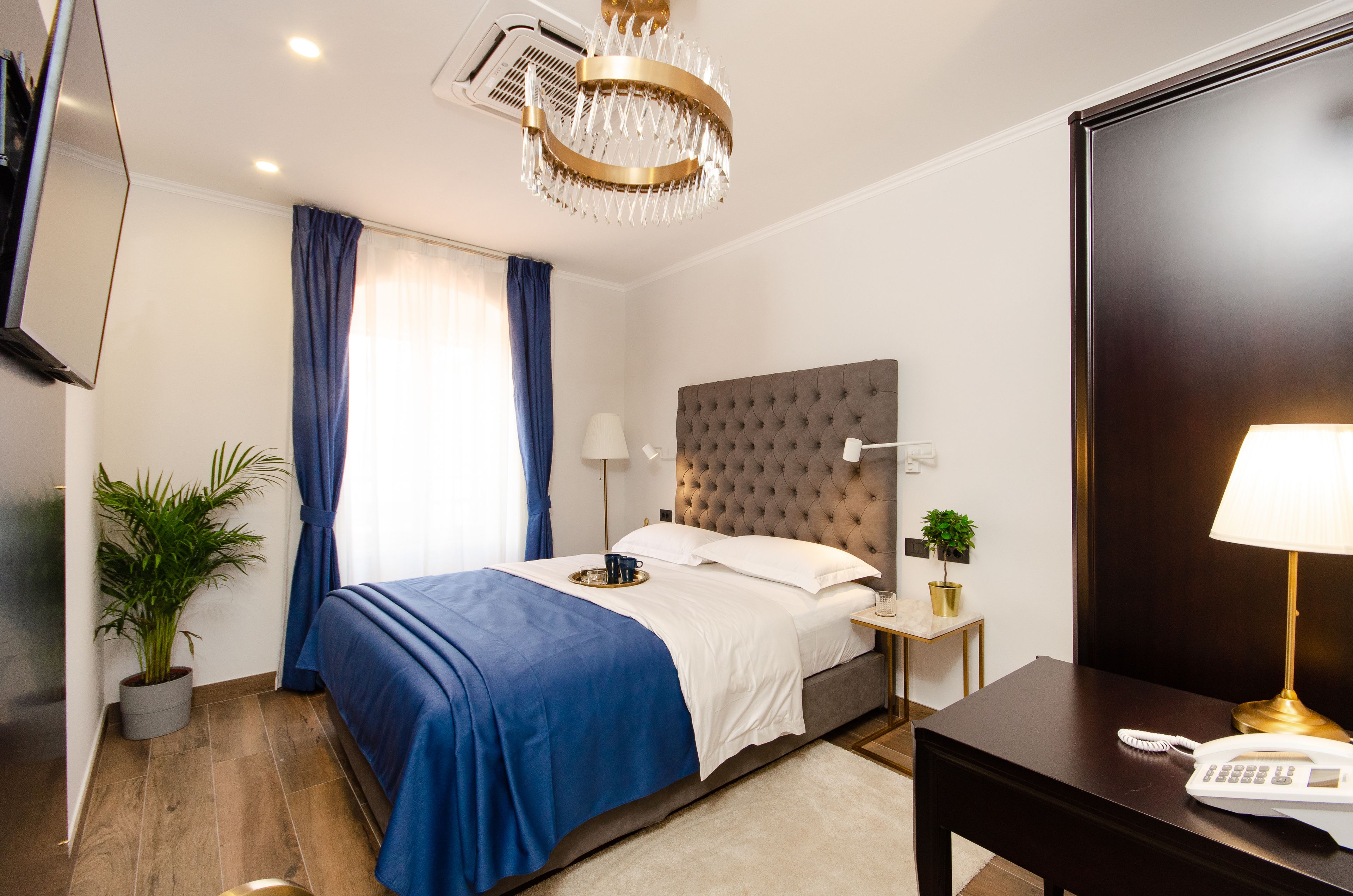 Habitación de hotel ordenada con cama, mesitas de noche, un escritorio con lámpara, televisor y planta en maceta.